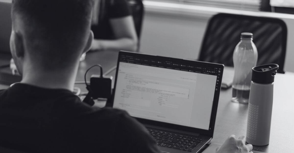 Homem digitando em um notebook que está em uma mesa. Ao lado do notebook há duas garrafas de água.